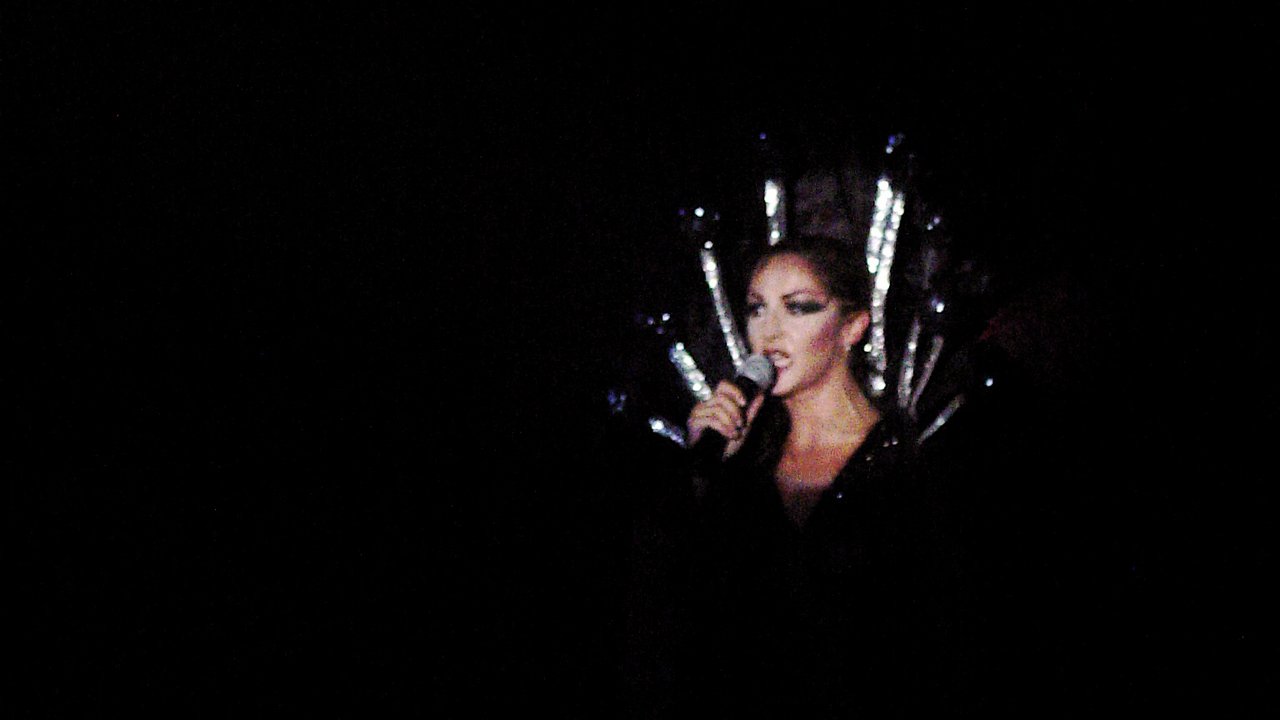 A performer in a silver spiked headdress singing into a microphone, surrounded by darkness