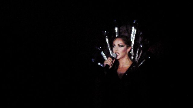 A performer in a silver spiked headdress singing into a microphone, surrounded by darkness