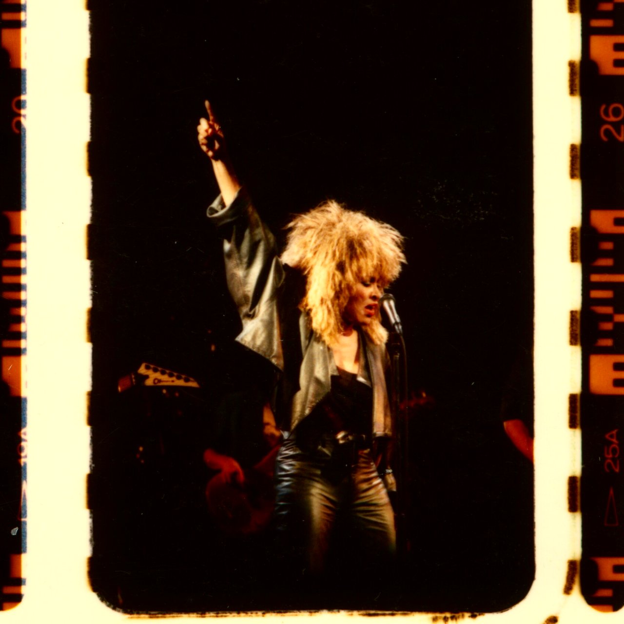 Tina Turner on stage in leather, fist raised — a 35mm film frame with sprocket holes and edge markings