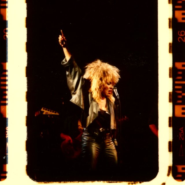 Tina Turner on stage in leather, fist raised — a 35mm film frame with sprocket holes and edge markings